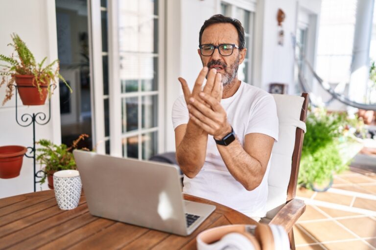 Middle age man using computer laptop at home suffering pain on hands and fingers, arthritis inflammation