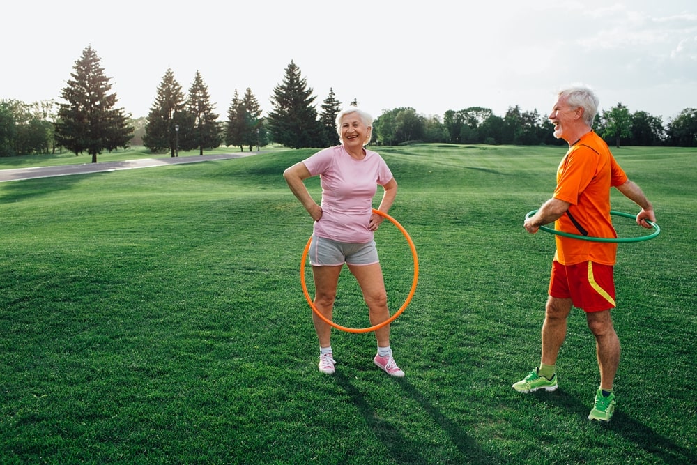 Elderly couple practices hula hoop exercises together on a grassy park area, enjoying the sunshine and staying active outdoors.